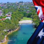 A Scenic Aerial View of An Australian Coastal Suburb with Lush Greenery, a Small Beach, and Houses, Accompanied by A Close-Up of The Australian Flag Waving in The Foreground