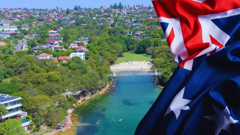 A Scenic Aerial View of An Australian Coastal Suburb with Lush Greenery, a Small Beach, and Houses, Accompanied by A Close-Up of The Australian Flag Waving in The Foreground