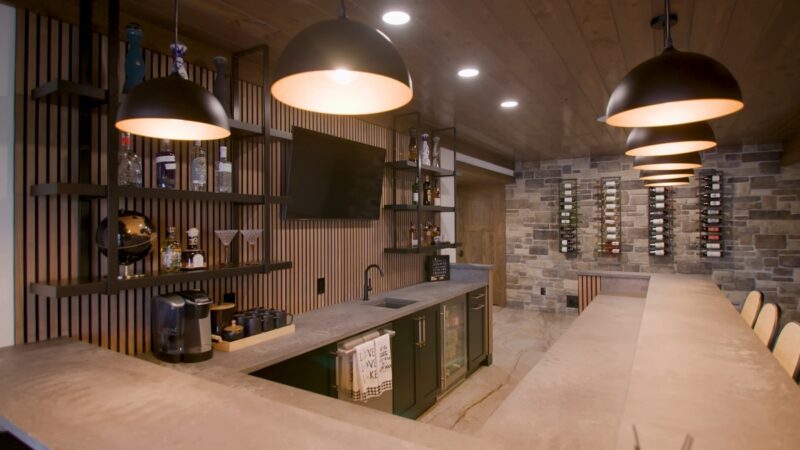 A stylish home bar with black pendant lights, a concrete countertop, a built-in sink, and shelves stocked with bottles and glasses