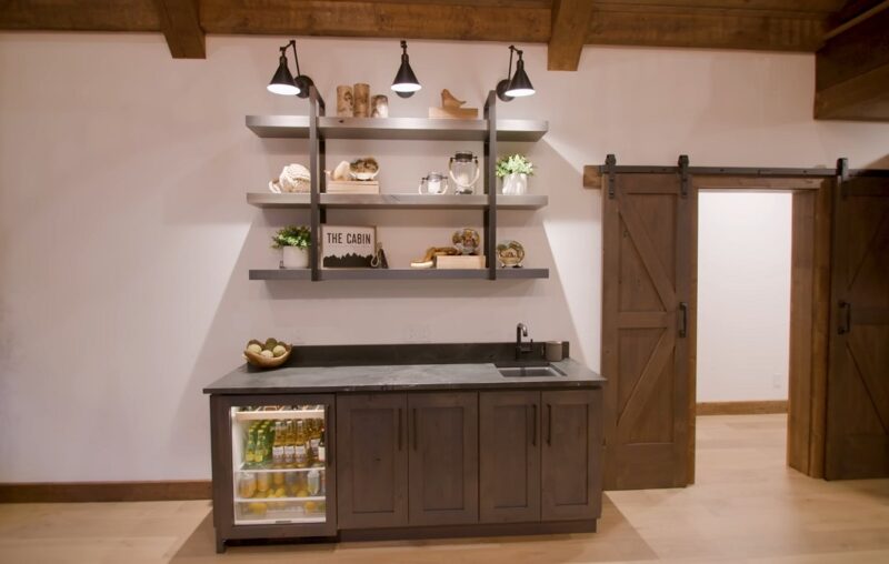 A stylish cabin bar area with dark cabinetry, floating shelves, a mini fridge, and wooden barn doors