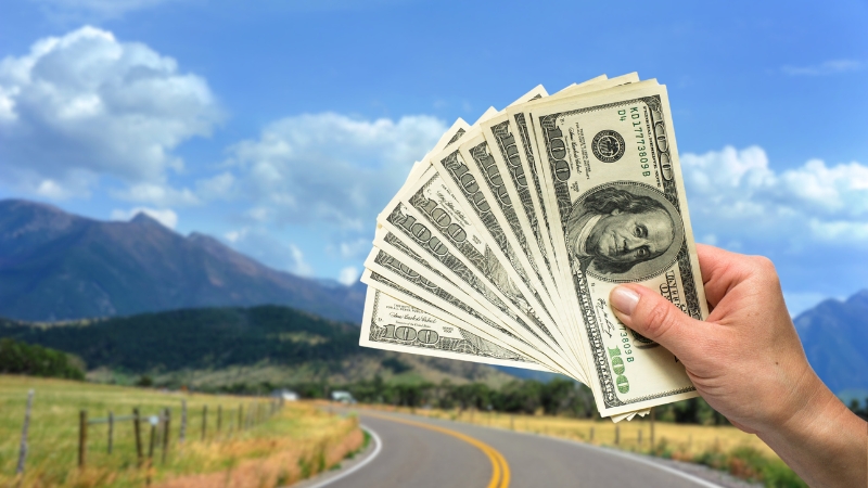 A Hand Holding Several $100 Bills Against a Scenic Montana Road Backdrop